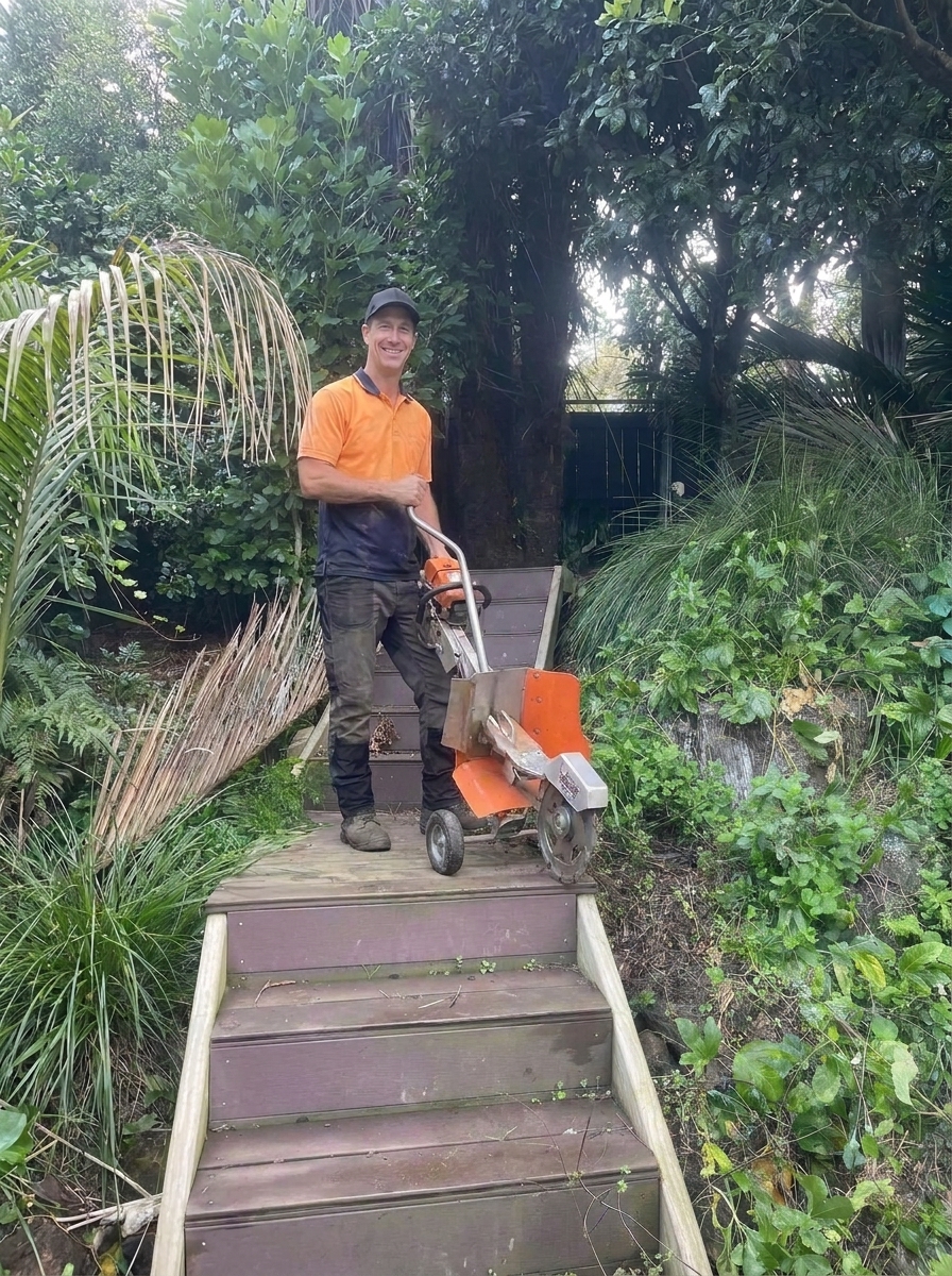 Operator taking a narrow-access stump grinder down bush steps to a rear North Shore garden
